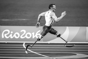 Ein Mann mit einer Prothese rennt auf einer Laufbahn mit einer Tafel im Hintergrund, die "Rio 2016 Paralympische Spiele" anzeigt, in Schwarz-Weiß.