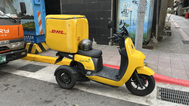 Gelber Elektro-Scooter auf einer Straße neben einem Lastwagen mit einem Helm darauf geparkt, Gebäude im Hintergrund und Menschen auf dem Gehweg.