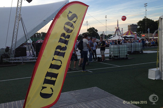 Ein buntes Karnevalsbild mit Zelten, einem zentralen Mast, Menschen, Bäumen, Strommasten, Straßenbahnen auf der Straße und einem eisernen Stand auf der linken Seite unter einem klaren Himmel.