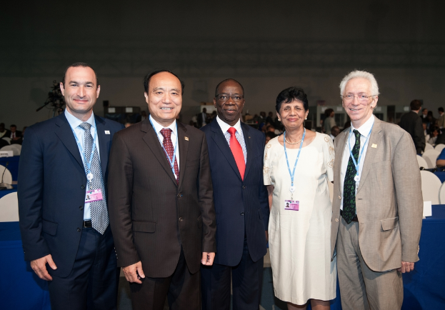 Eine Gruppe von Männern in Anzügen und eine Frau in einem Kleid steht und lächelt in der Nähe einer Halle, mit Menschen, die auf Stühlen und Tischen sitzen und stehen, einem Mikrofon auf einem Tisch links und einer Wand und anderen Gegenständen im Hintergrund.