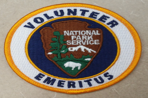 Ein runder Fleck mit einem blauen Rand, einem weißen Hintergrund und einer fetten schwarzen Schrift, die den Text "Volunteer National Park Service Emeritus" in der Mitte eines gelben Kreises zeigt.