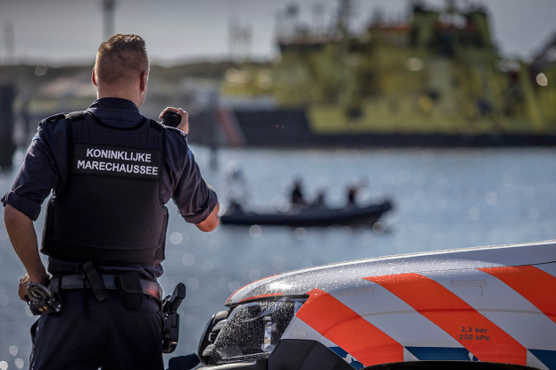 Ein Polizist in Uniform steht neben einem Polizeiwagen in der Nähe eines Gewässers und hält ein Funkgerät in der Hand, im Hintergrund ist ein Boot mit ein paar Leuten auf dem Wasser zu sehen und der Himmel darüber.