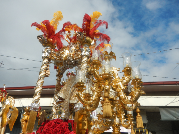 Ein prächtiger goldener und roter Wagen, geschmückt mit Blumen und anderen Dekorationen bei einem Karnevalsumzug, mit einem Gebäude, Strommasten mit Drähten und einem bewölkten Himmel im Hintergrund.