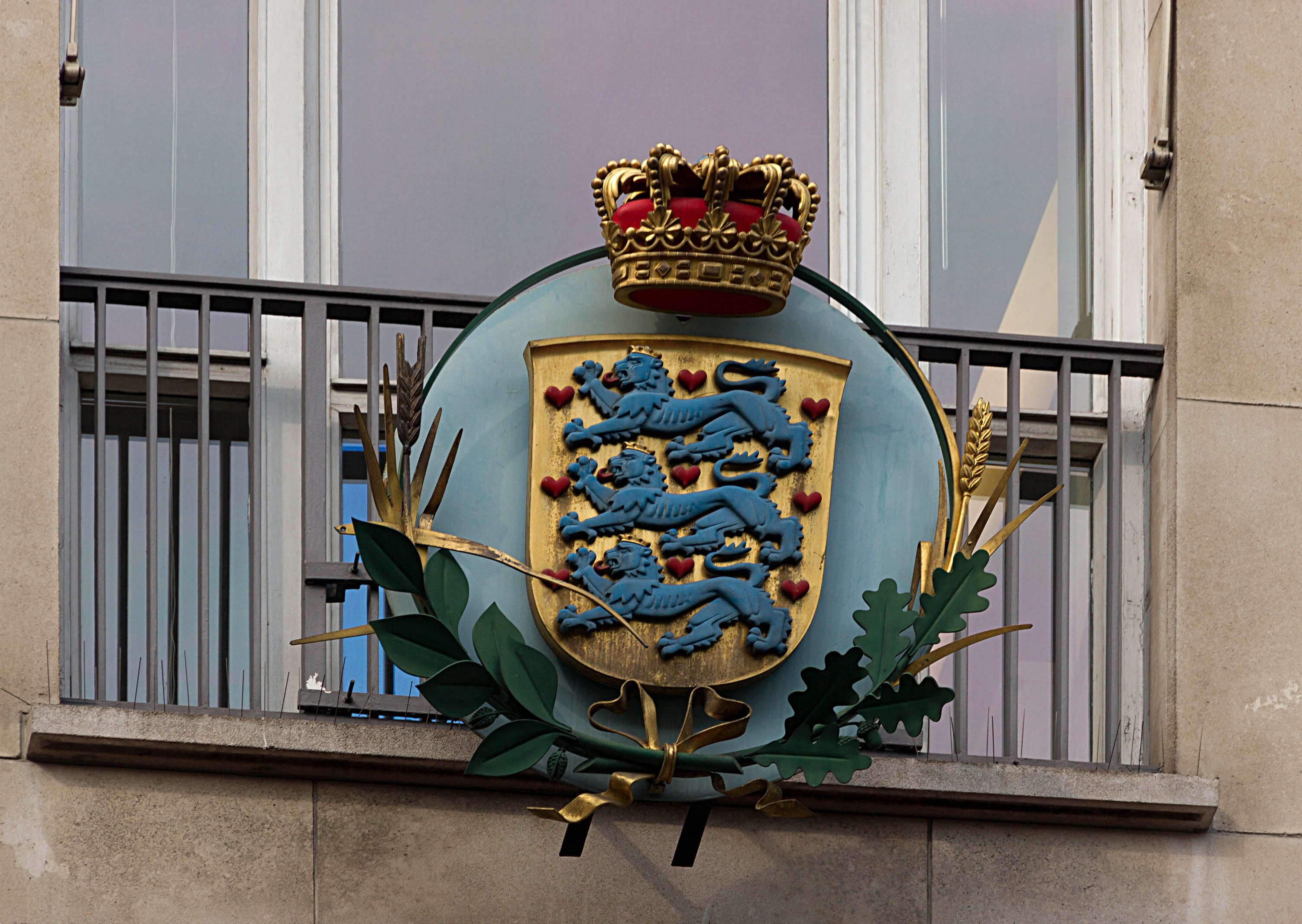 Wappen der britischen Königsfamilie, das auf der Außenseite eines Gebäudes angezeigt wird und ein Logo mit Krone und Blatt zeigt, mit Fenstern und Metallgittern im Hintergrund.