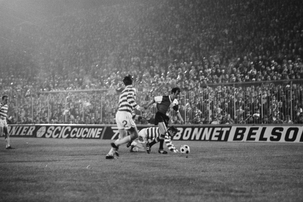 Eine Gruppe von Männer, die auf einem Fussballfeld Fussball spielen, mit einer Zuschauermenge auf den Tribünen und einem Banner mit der Aufschrift "Celtic v West Bromwich Albion - FA Cup Third Round" in Schwarz-Weiß.