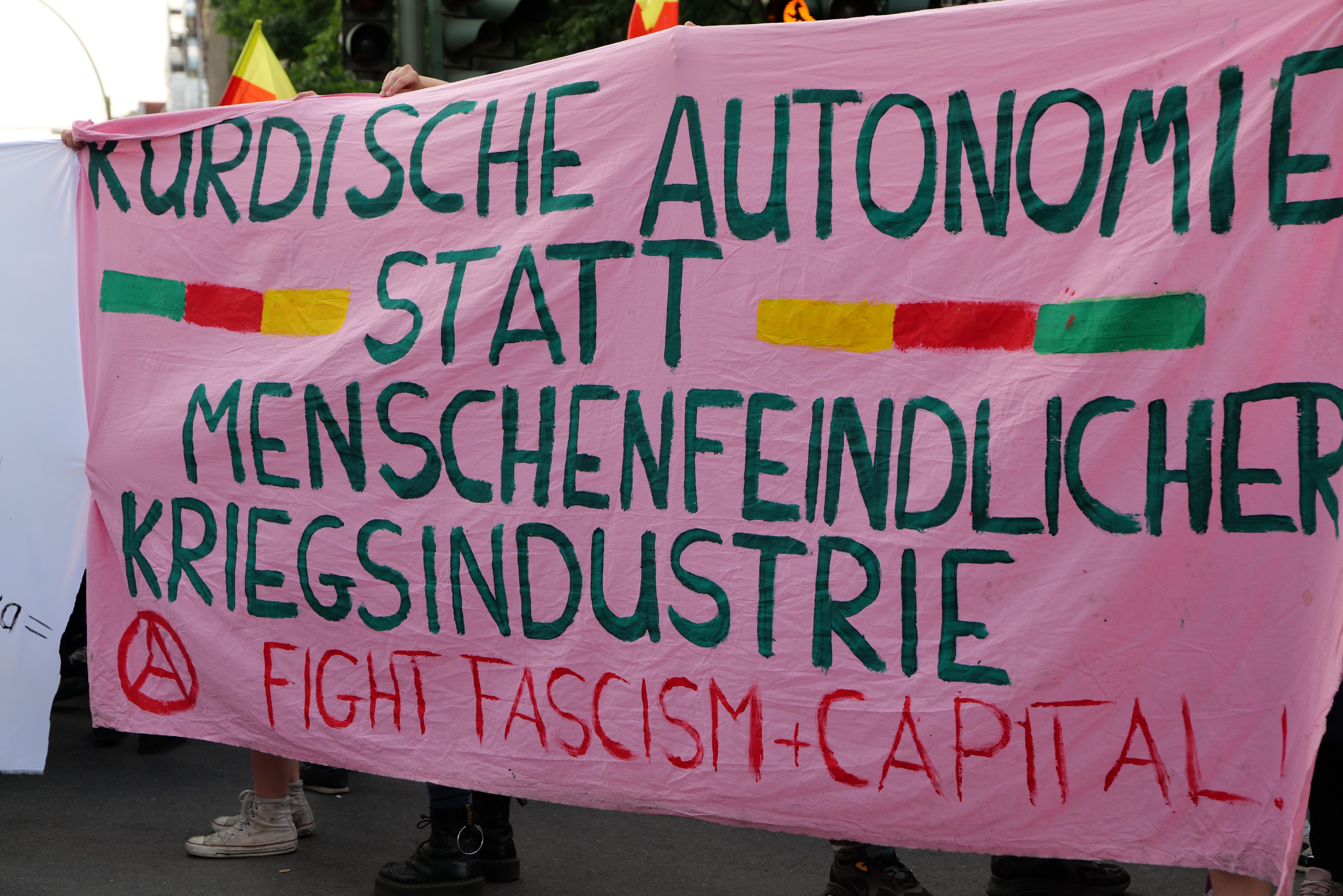 Eine Gruppe von Menschen mit einer pinken Fahne, auf der "Kurdische Autonomie Statt Menschenfeindlicher Kriegsindustrie Fight Fascism Capital" steht, auf einer Straße mit Ampeln, Flaggen, Bäumen, Gebäuden und einem klaren blauen Himmel im Hintergrund.