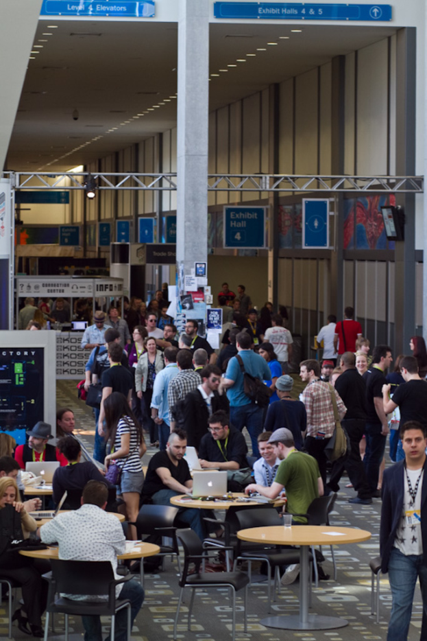 Ein belebter Flughafenvorplatz mit wartenden Menschen, einige stehen und andere arbeiten, Richtungsschilder an der rechten Wand und Ã¼ber ihnen, und Lichter, die von der Decke hÃ¤ngen.