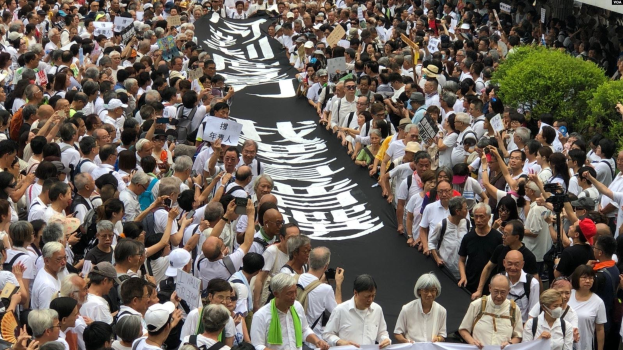 Große Gruppe von Menschen marschiert durch eine Straße, hält Protestschilder und Banner in der Hand, mit Grünzeug auf der rechten Seite und einem Gebäude im Hintergrund.
