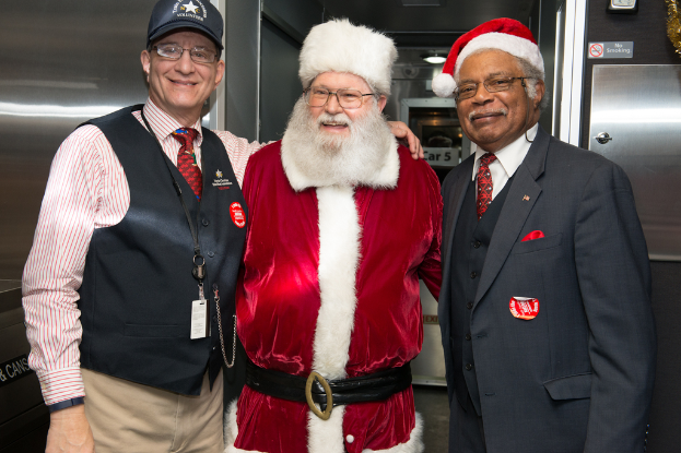 Eine Gruppe von Menschen in Brillen, Mützen, Krawatten, ID-Karten und Abzeichen, mit einer Person in einem Weihnachtskostüm, die vor einer Stahlwand mit Lichtern, Schildern und anderen Gegenständen im Hintergrund steht.