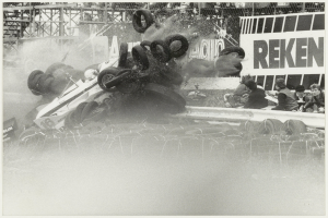 Ein Schwarz-Weiß-Foto eines zertrümmerten Rennwagens umgeben von Reifen, mit einer Gruppe von Menschen rechts und einer Tafel mit Text im Hintergrund.