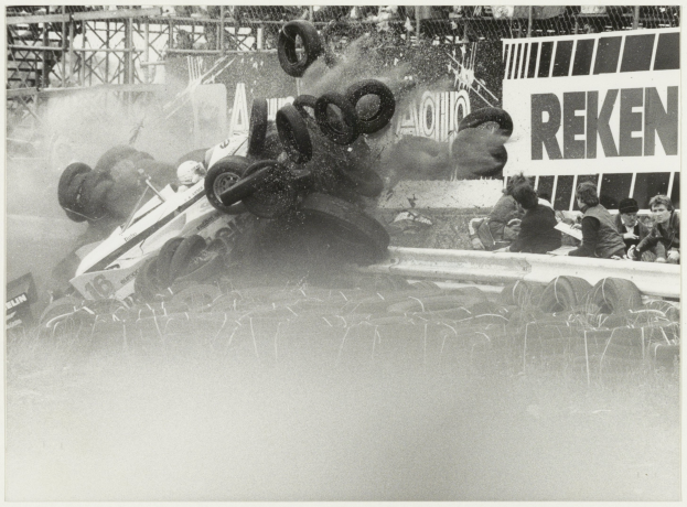 Ein Schwarz-Weiß-Foto eines zertrümmerten Rennwagens umgeben von Reifen, mit einer Gruppe von Menschen rechts und einer Tafel mit Text im Hintergrund.