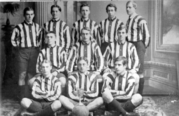 Ein Schwarz-Weiß-Foto einer Fußballmannschaft, mit einigen stehenden und anderen auf dem Boden sitzenden Spielern, einem Pokal in der Mitte, Säulen und einer Wand im Hintergrund und Text oben und unten auf dem Foto.