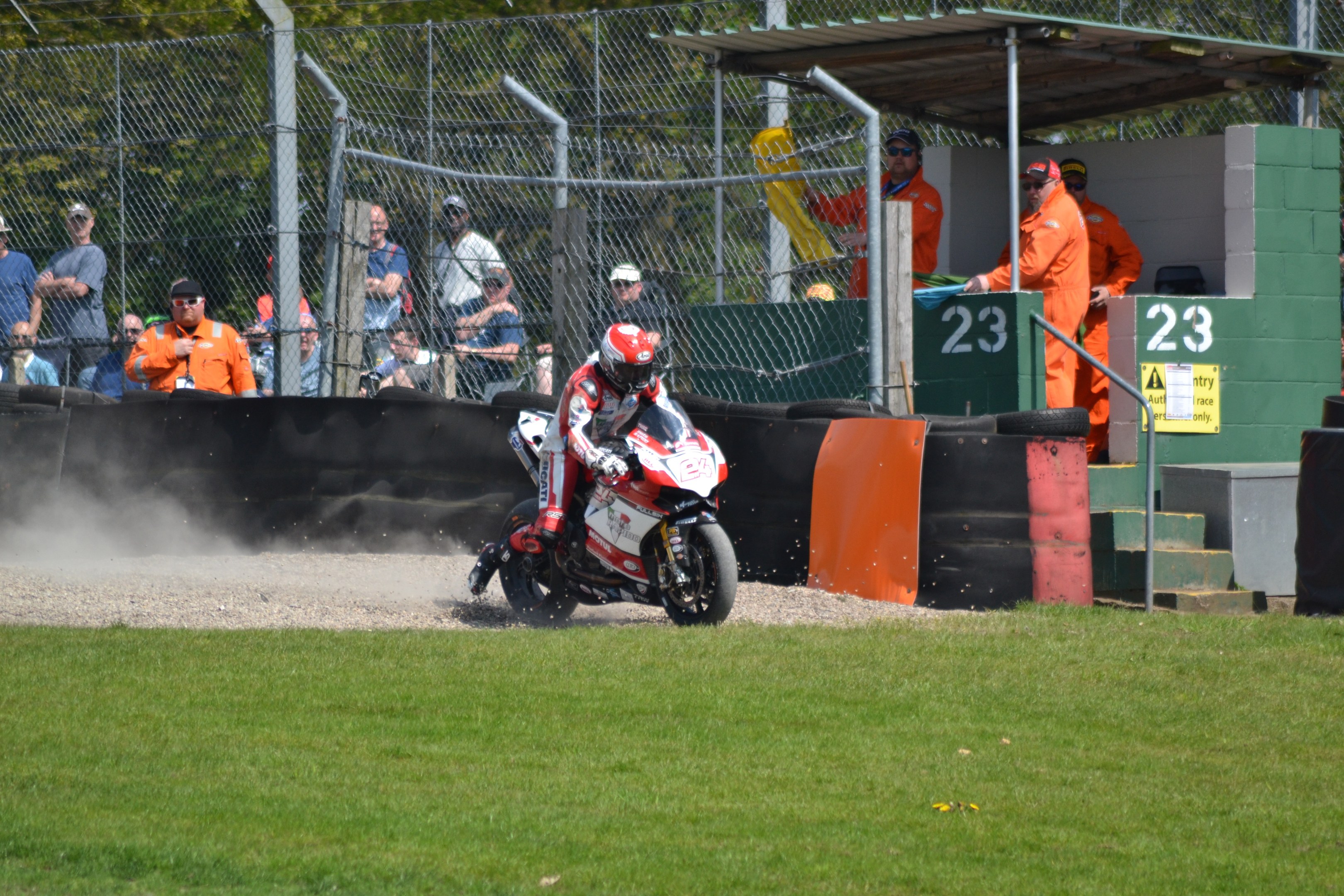 Eine Person in Helm und Handschuhen fährt ein Motorrad über ein grasbewachsenes Feld, mit Zuschauern in Mützen und Schutzbrillen, einem eingezäunten Bereich, Treppen mit Geländern und einem nummerierten Schuppen im Hintergrund.