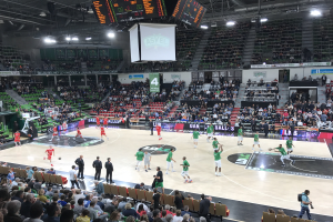 Eine Gruppe von Menschen, die Basketball in einer vollen Arena mit Zuschauern in den Rängen spielt, beleuchtet von Stadionlampen und Bildschirmen.