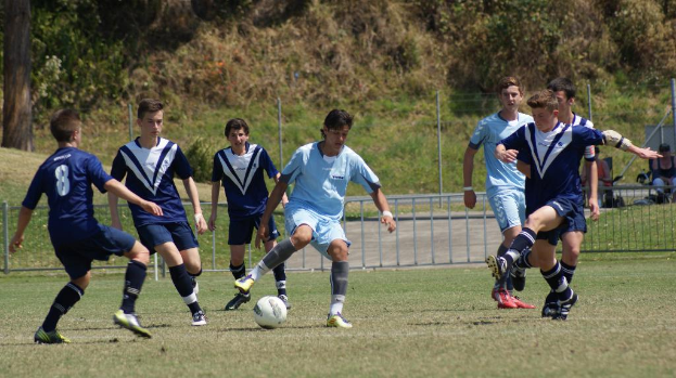 Eine Gruppe von Menschen spielt Fußball mit einem Ball auf dem Boden, während zwei Personen im Hintergrund auf Stühlen sitzen und weitere Bäume im Hintergrund zu sehen sind.