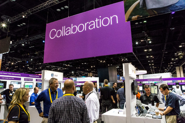 Eine Gruppe von Menschen, die stehen, einige halten ID-Karten, mit einem Mann, der an einem Laptop arbeitet, an einem Tisch, auf dem eine Tasse, ein Fernseher, ein Telefon und ein paar Laptops stehen; es gibt Werbetafeln und Deckenleuchten im Hintergrund.