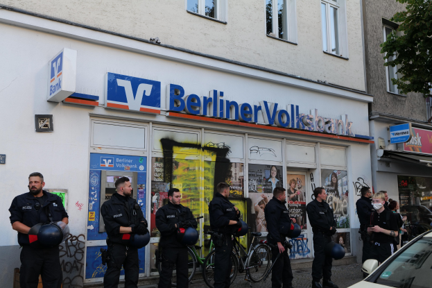 Gruppe von Polizisten vor einer Berliner Volksbank stehend, einige halten Helme, mit einem Auto rechts und einem Baum links, Gebäude und Texttafeln im Hintergrund.