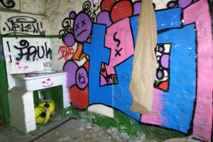 Ein beschmierter Badezimmer mit einem mit einem Tuch bedeckten Waschbecken und verstreuten Gegenständen auf dem Boden.