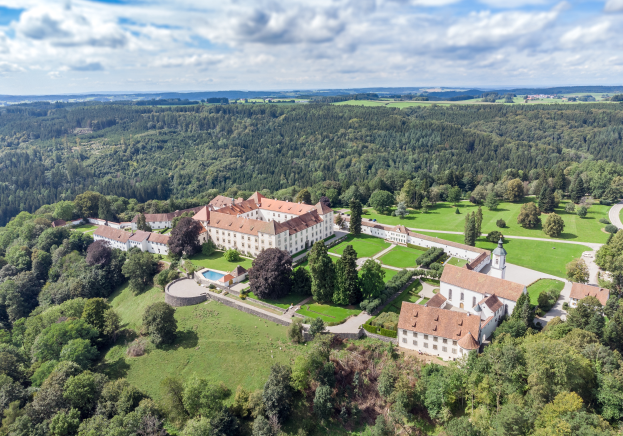 Luftaufnahme eines großen Schlosses in einem Wald, umgeben von grünem Gras, Bäumen und Pflanzen, unter einem Himmel voller weißer, flauschiger Wolken in der bayerischen Landschaft.