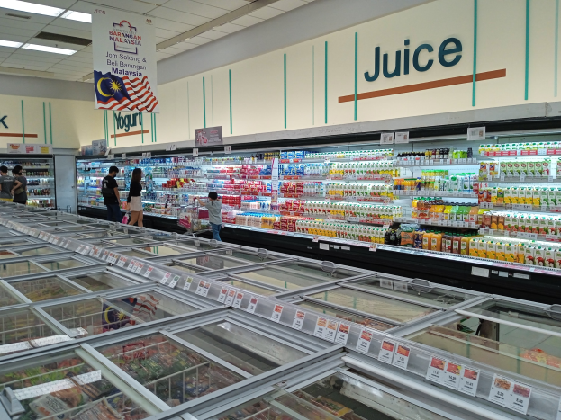 Ein hell erleuchteter Saftladen in einem Supermarkt zeigt eine Reihe von Flaschengetränken, frischem Gemüse und anderen Artikeln auf Regalen, mit einigen Kunden, die durch die Gänge schlendern und Deckenleuchten, die die Szene beleuchten.