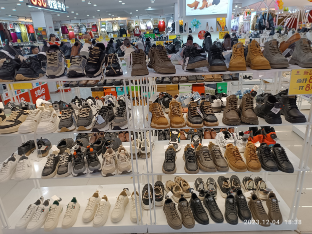 Ein Schuhgeschäfts-Innenraum mit mehreren Regalen voller Schuhe, jeweils mit Preisschildern, Menschen im Hintergrund, hängende Kleidung, Deckenleuchten, ein Bildschirm und Informationsschilder.