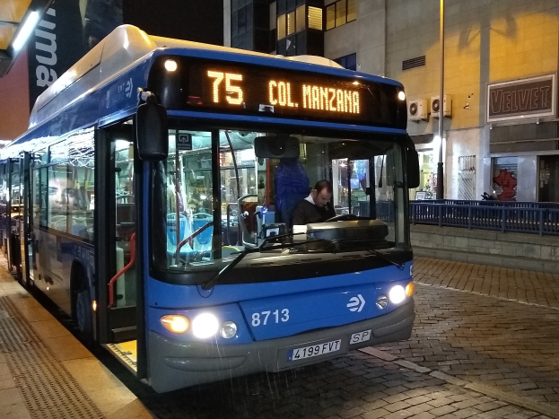 Ein blauer Bus parkt nachts am Straßenrand mit einer Person darin, einer Handtasche im Vordergrund und Gebäuden, Schildern, Geländern, Pfählen und Laternen im Hintergrund.
