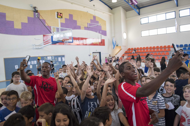 Kinder vor einem Basketballfeld an einem Camp, einige halten Telefone, mit einem Korb, Netz, Uhr, Papieren an einer Tafel, Deckenlichtern, Stühlen und Fenstern im Hintergrund.