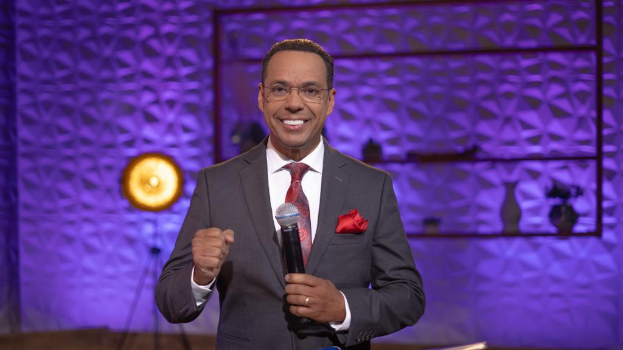 Stephen Colbert, der eine Anzug, Krawatte und Brille trägt, steht auf einer Bühne und hält ein Mikrofon in der Hand, mit einem Ständer und Licht hinter ihm.