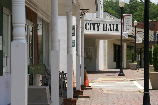 Ein Rathaus umgeben von Gebäuden, Straßenmasten, Straßenlaternen, Namensschildern, Kraftfahrzeugen, Verkehrskegeln, Stühlen, Topfpflanzen, Bäumen und einer bewölkten Himmel
