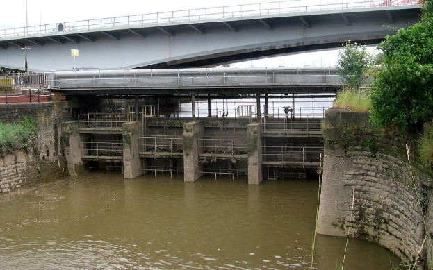Ein Blick auf die Schleusentore mit fließendem Wasser, eine Brücke und umliegende Bäume und Pflanzen.