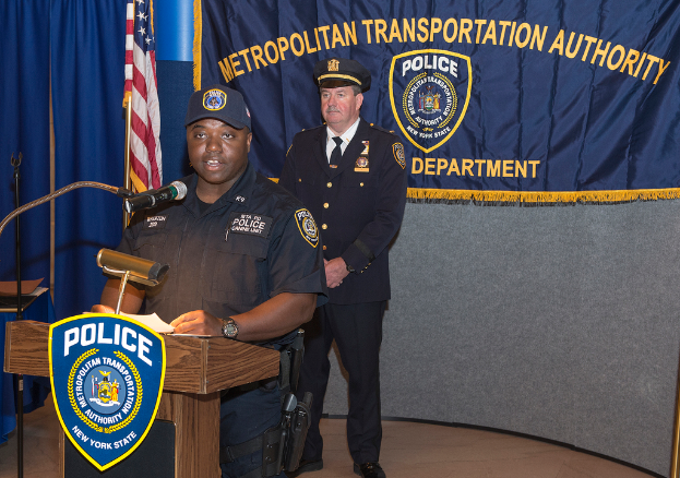 Zwei Polizisten stehen vor einem Rednerpult, einer hält Papiere und spricht in ein Mikrofon, mit einer Tafel, Flagge und Tisch in der Nähe und einer Tafel mit Symbolen und Text im Hintergrund.