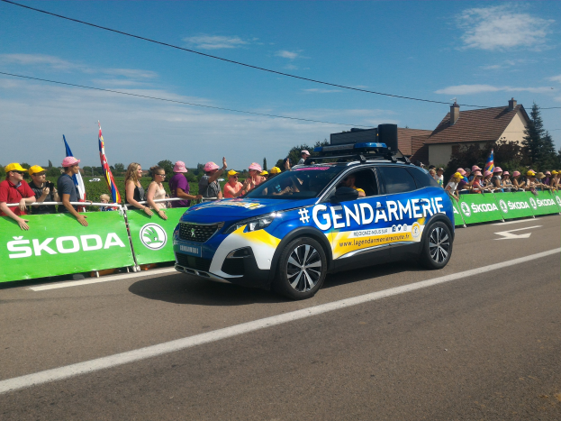 Polizeifahrzeug fährt neben einer Menschenmenge mit Fahnen, mit einem Banner auf dem Auto und Gebäuden im Hintergrund, beschriftet mit "Gendarmerie de la Tour de France."