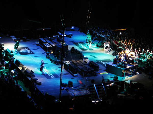 Eine Gruppe von Menschen, die auf einer Bühne mit Lautsprechern, Scheinwerfern und Mikrofonen performen, umgeben von einer großen Menge in einer dunklen Umgebung.