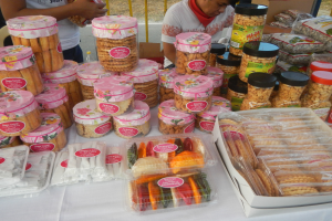 Eine Gruppe von Menschen steht um einen Tisch herum, der mit verschiedenen Lebensmitteln, darunter Schachteln und Packungen, gefüllt ist, mit einem Geländer und einer Straße im Hintergrund auf einem Food-Festival.