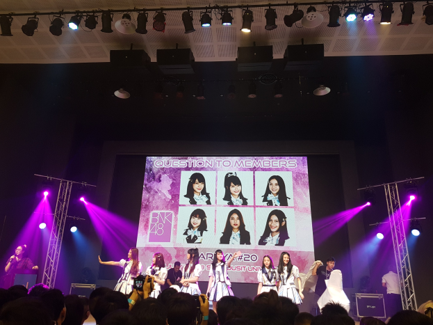 Eine Gruppe von Menschen, die auf einer hell erleuchteten Bühne performen, mit einer Menge darunter, einem Sänger mit Mikrofon und einem Musiker, der ein Instrument spielt, während ein großes Display Bilder der K-Pop-Gruppe BK48 zeigt.