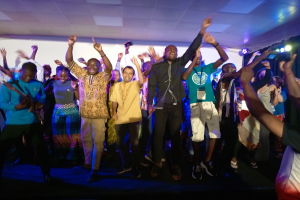 Eine Gruppe von Menschen, die auf einer Bühne tanzen und ihre Hände in die Luft strecken, beleuchtet von Scheinwerfern auf der rechten Seite und einem Bildschirm im Hintergrund, unter einer Decke mit Scheinwerfern.
