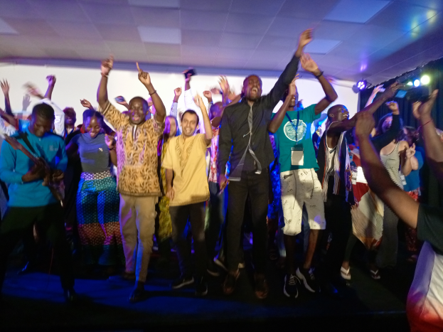 Eine Gruppe von Menschen, die auf einer Bühne tanzen und ihre Hände in die Luft strecken, beleuchtet von Scheinwerfern auf der rechten Seite und einem Bildschirm im Hintergrund, unter einer Decke mit Scheinwerfern.