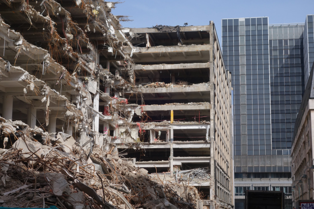 Ein großes Gebäude in einer Stadt abbruchreif, umgeben von intakten Strukturen, Straßeninfrastruktur, Fahrzeugen, Bäumen und einem klaren Himmel.