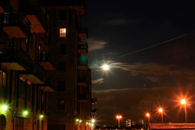 Eine nächtliche Stadtansicht mit einem prominenten Gebäude mit zahlreichen Fenstern und Balkonen im Vordergrund und mehreren Straßenlaternen auf der rechten Seite.