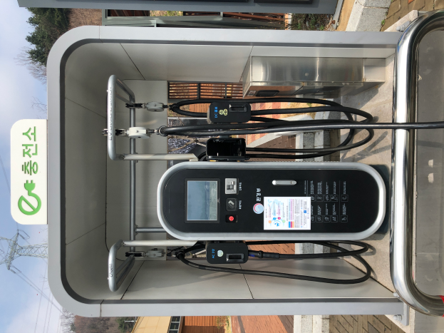 Eine Ladestation für Elektrofahrzeuge mit einem Gerät, Kabeln und einer Texttafel vor dem Hintergrund von Gebäuden, Bäumen, Masten, Kabeln und dem Himmel in Seoul, Südkorea.