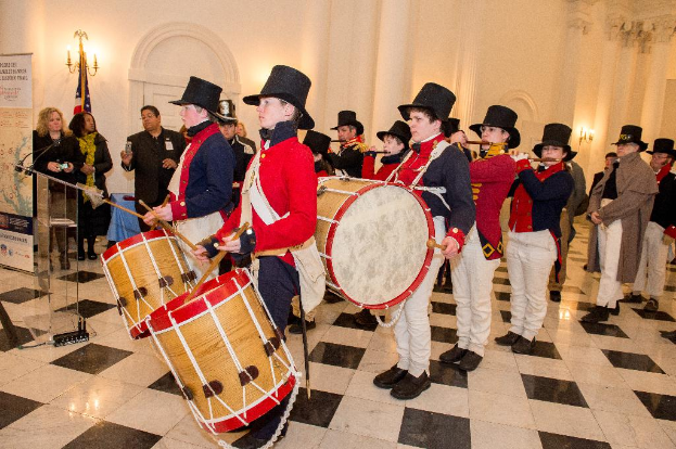 Eine Gruppe von Musikern, die auf der Bühne in einem Saal mit strukturierten Wänden Trommeln und Flöten spielt, während sich Menschen um sie sammeln.