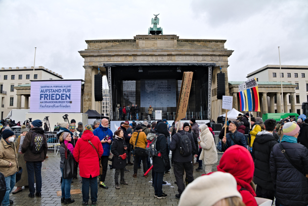 Eine Menschenmenge steht vor einem Gebäude mit einer Bühne, auf der Redner und ein Bildschirm zu sehen sind, flankiert von Fahnen und Transparenten mit Text auf der rechten Seite, in einer Protestszene in Berlin mit bewölktem Himmel und Gebäuden im Hintergrund.