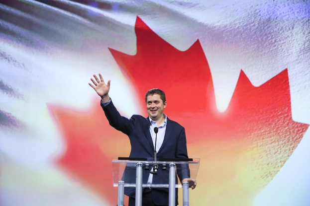 Der kanadische Premierminister Alex Salmond steht an einem Rednerpult mit Mikrofonen, lächelt vor einer kanadischen Flagge, wobei ein Bildschirm im Hintergrund sichtbar ist.