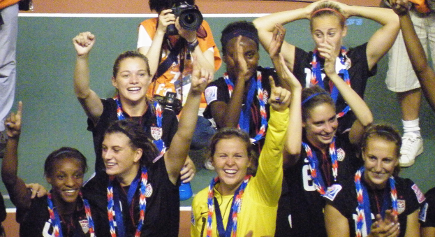 Eine Gruppe von Frauenfußballspielerinnen auf einem Feld stehend, lachend und mit Medaillen behängt, im Hintergrund ein Mann mit einer Kamera.