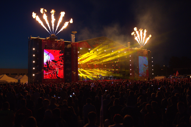 Eine große Menschenmenge auf einem Musikfestival mit einer beleuchteten Bühne, die von Performern umgeben ist, links von Zelten und einem dunklen Himmel mit Feuerwerk.