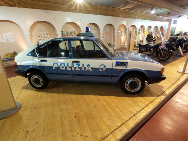 Ein Polizeiwagen wird in einem Museum ausgestellt, umgeben von Motorrädern und anderen Fahrzeugen, mit einer Säule und einer Tafel auf der linken Seite, Plakaten an den Wänden und einer Beleuchtung von der Decke.