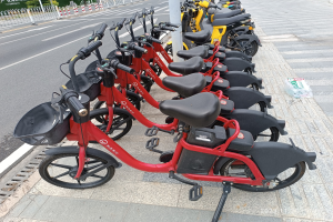 A row of red and black e-bikes parked along a street with a white pole in the foreground, trees, buildings, and a clear blue sky in the background.