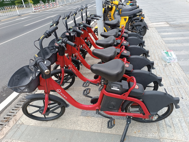 A row of red and black e-bikes parked along a street with a white pole in the foreground, trees, buildings, and a clear blue sky in the background.