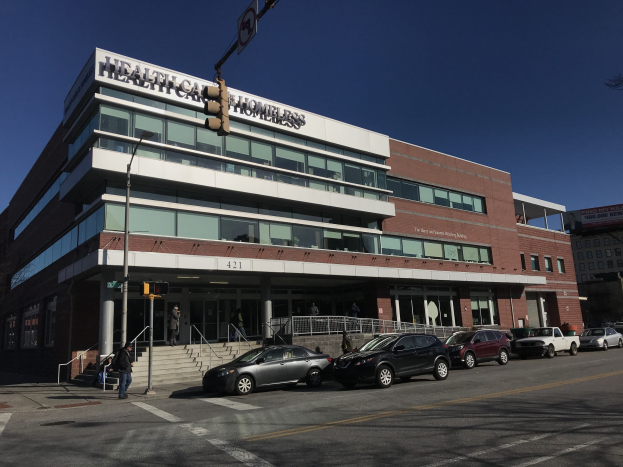 Großes Gebäude mit der Aufschrift 'New York City Health Care Homeless Center' mit parkenden Autos, Fußgängern, Straßenmöbeln und Bäumen unter einem klaren Himmel.