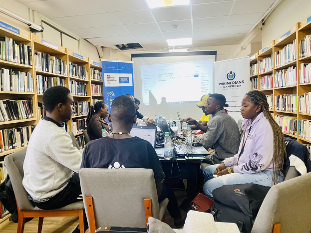 Eine Gruppe von Menschen, die um einen Tisch in einer Bibliothek sitzen, umgeben von Bücherregalen, mit Laptops, Flaschen und Taschen auf und um den Tisch, diskutieren eine Wikimedians und Kamerun-Initiative.